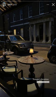 two chairs and a table on the side of the road at night with cars parked in the background