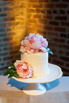 a white cake with pink flowers on top sits on a table in front of a brick wall