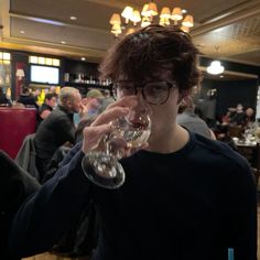 a young man drinking from a wine glass in a crowded restaurant with people sitting at tables