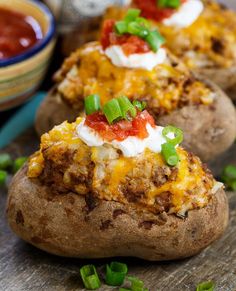 three loaded baked potatoes on a cutting board with green onions and salsa in the background