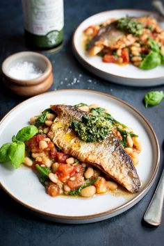 two white plates topped with fish and beans next to a bottle of wine on a table