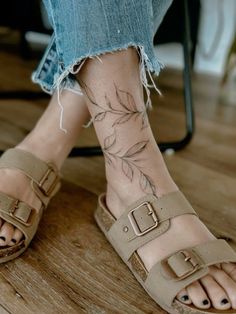 a woman's legs with tattoos on them and her feet in sandals, sitting on a wooden floor