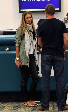 a man and woman standing in front of a tv screen at an airport terminal, talking to each other