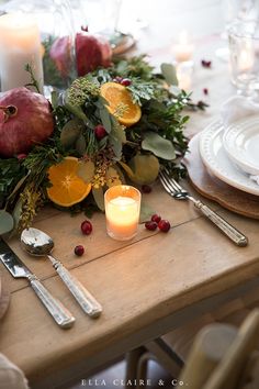 an arrangement of fruit and greenery on a table with silverware, candles and plates