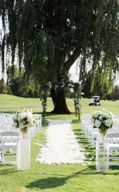 an outdoor ceremony setup with white chairs and flowers on the aisle, in front of a large tree