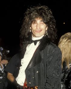 a man with long hair wearing a black jacket and tie standing next to other people