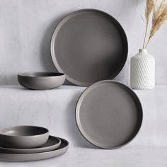 a set of grey dinnerware on a white marble countertop next to a vase with dry grass in it