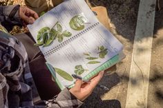 a person is holding a notebook with plants on it and writing down the page in front of them