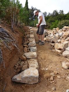 a man standing next to a pile of rocks on top of a dirt road with trees in the background