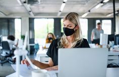 a woman wearing a face mask while working on her laptop in an office with other people