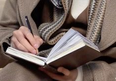 a person sitting down and writing on a book with a pen in their hand while wearing a coat