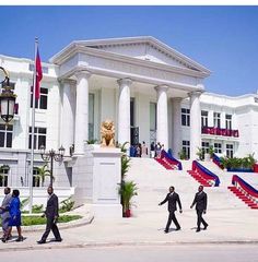 several people walking in front of a building with columns and flags on the side walk