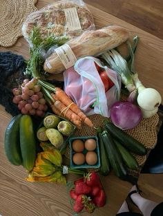 an assortment of fruits and vegetables sitting on a wooden table next to bread, eggs, tomatoes, cucumbers, carrots, broccoli