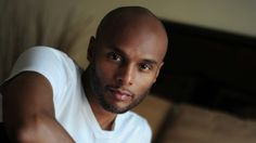 a bald man in a white t - shirt sitting on a bed looking at the camera