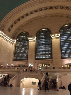 people are standing in the lobby of a train station with large windows and lights above them