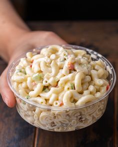 a person holding a plastic bowl filled with macaroni and cheese salad on top of a wooden table