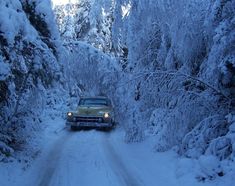 an old car driving down a snow covered road in the middle of trees and bushes