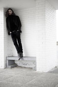 a man with long hair and beard leaning against a white brick wall on a bench