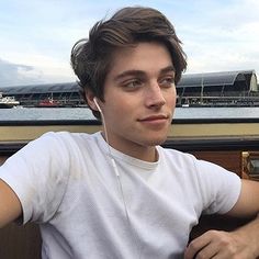 a young man sitting on top of a wooden bench next to the ocean with headphones in his ears