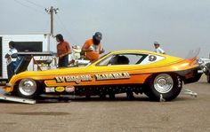 an orange car is being towed by two men in front of a trailer with other cars behind it