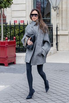 a woman in grey coat and black tights walking down the street with her hand on her hip