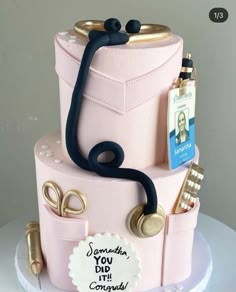 a pink cake with gold accents and writing on the top is adorned with a doctor's stethoscope