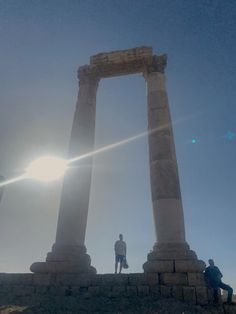 two people standing on top of a stone wall next to large pillars with the sun shining through them