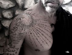 a man with tattoos on his chest standing next to a stone wall and looking at the camera