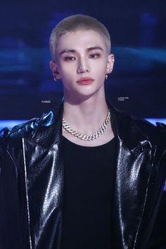 a young man wearing a black leather jacket and silver necklace, standing in front of a blue background