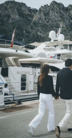 two people are walking in front of some yachts and one is holding the hand of another person's hand