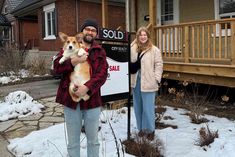 a man and woman standing in front of a sold sign with a dog on his lap