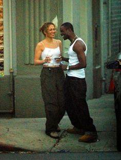 a man and woman standing on the sidewalk talking to each other while holding drinks in their hands