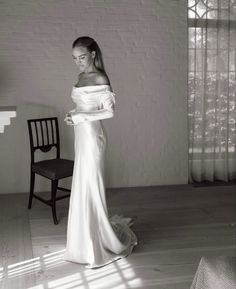 a woman in a long white dress standing next to a chair and looking off into the distance