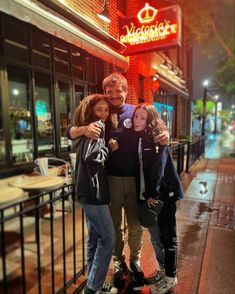 three people posing for a photo in front of a restaurant on a rainy night with their arms around each other