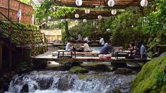 people sitting at tables near a waterfall in the forest with lanterns hanging from the roof