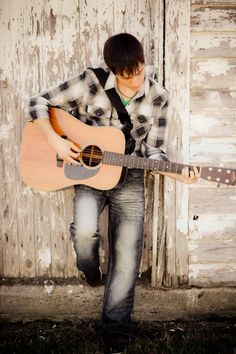 a young man is holding an acoustic guitar in front of a barn door and leaning against the wall