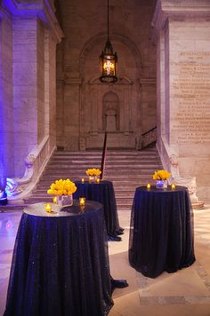 two tables with blue cloths and yellow flowers on them in front of a staircase