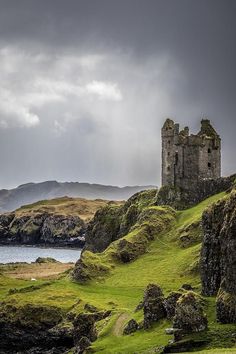 an old castle sitting on top of a lush green hillside next to the ocean under a cloudy sky