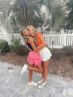 two young women hugging each other in front of a white fence and palm tree on the sidewalk