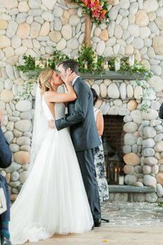 a bride and groom kissing in front of a stone fireplace at their outdoor wedding ceremony