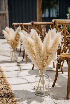 two clear vases filled with dried flowers on top of a stone floor next to wooden chairs