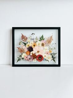 a black frame with flowers and leaves in it on a white wall next to a shelf