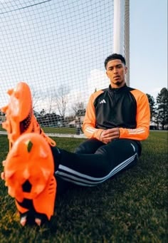 a man sitting on the ground in front of a soccer goal wearing an orange and black jacket