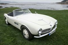 an old white sports car parked on the grass near water and rocks in the background