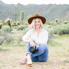 a woman sitting on the ground with a camera in her hand and wearing a hat