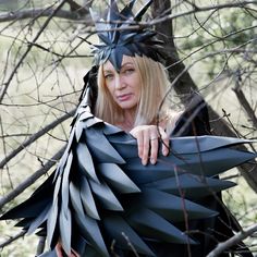 a woman wearing a black bird costume standing in the woods with her hands on her chest