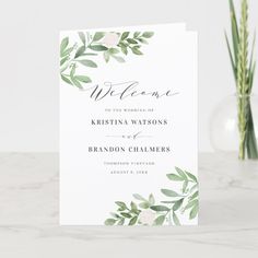 a wedding welcome card with greenery and white flowers on the front, sitting next to two vases