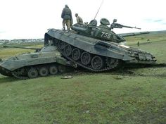 two men standing on top of a tank in the middle of a grassy field with other tanks