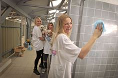 three women in white shirts are cleaning a tiled wall with a rag and a dog