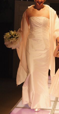 a woman in a white dress walking down a runway with a flower bouquet on her hand